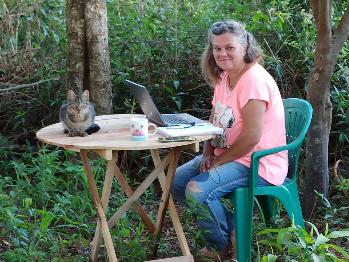 Andrea Marohn sitzt lächelnd an einem Holztisch im Grünen, mit Laptop, Tasse und Katze
