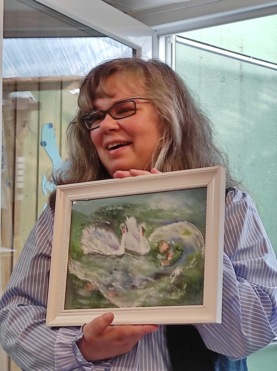 Andrea Marohn lacht und hält ihr Aquarell mit dem Schwanenpaar im hellen Holzrahmen